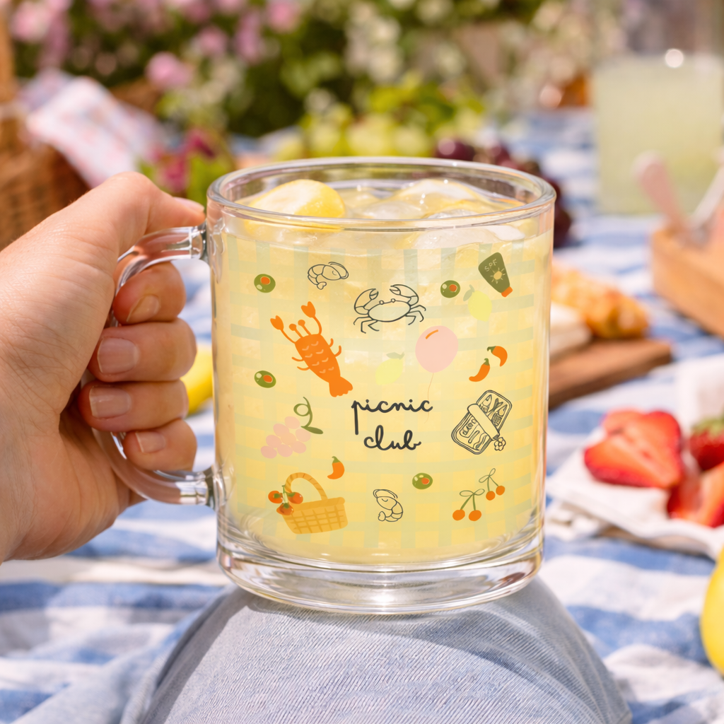 Caneca de vidro com ilustrações e líquido amarelo claro segurada numa mão em piquenique