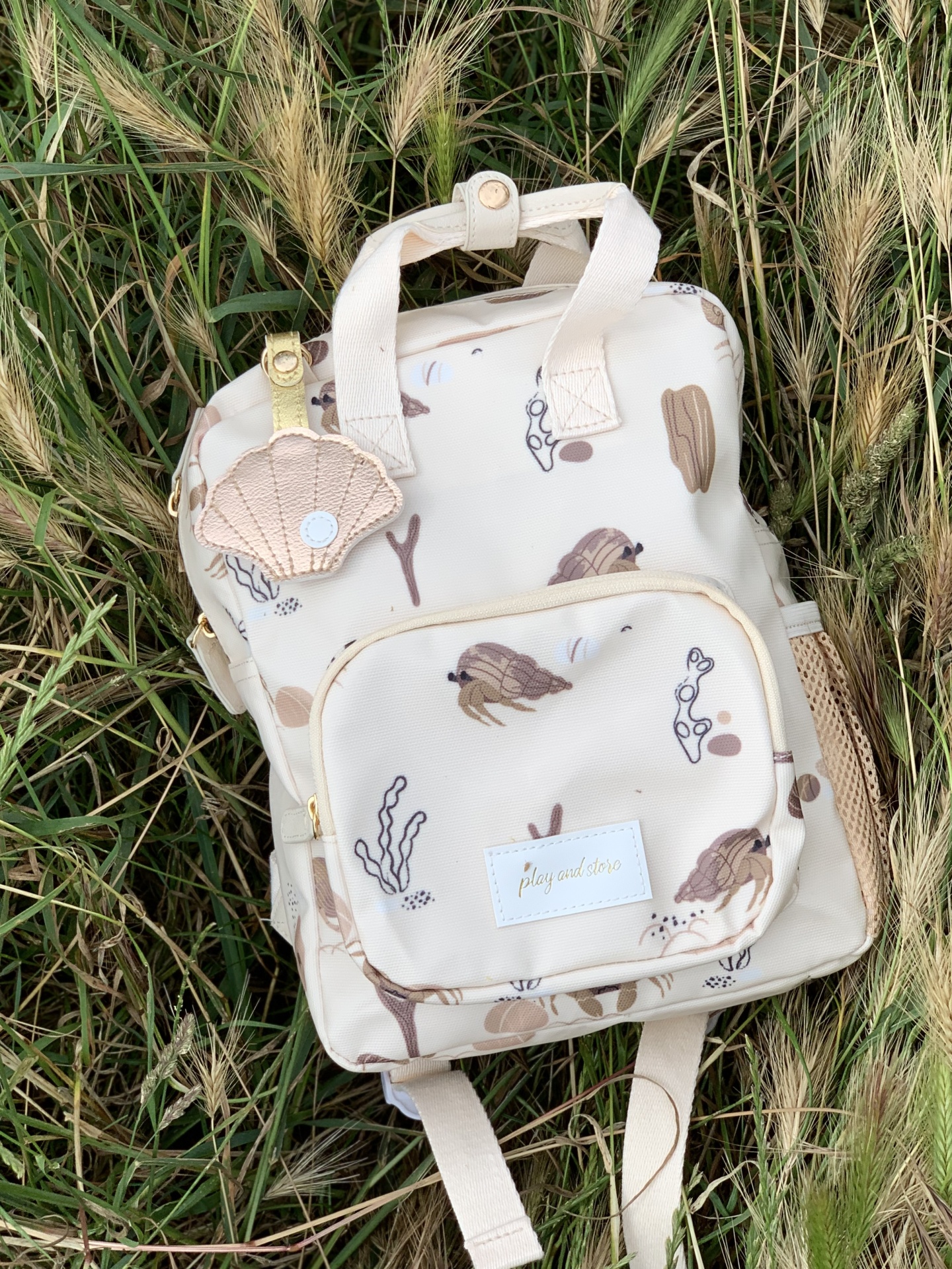 Mochila creme com padrão marinho e chaveiro em forma de concha em relva alta