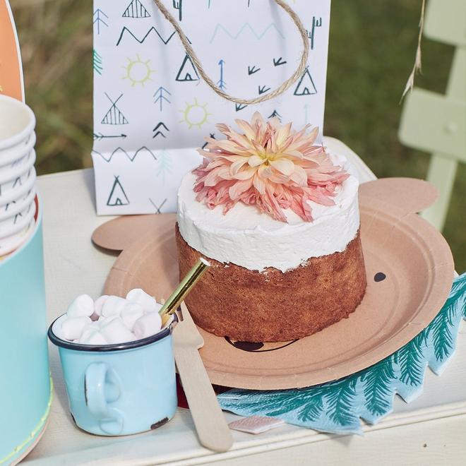 Bolo decorado com flor, prato de papel, caneca azul com marshmallows e guardanapo verde numa mesa clara