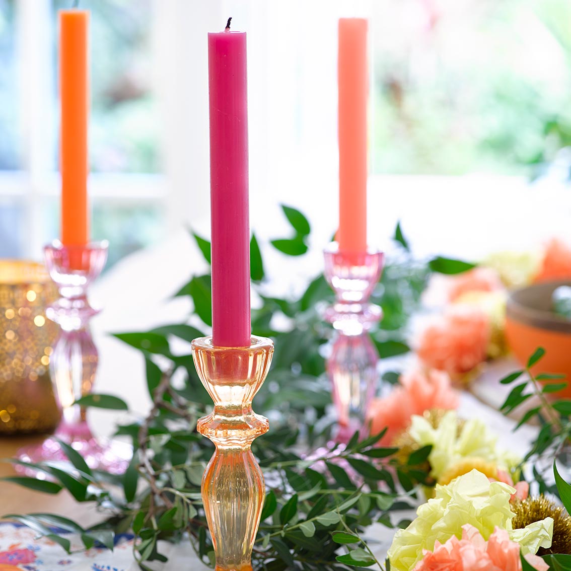 Castiçais de vidro com velas coloridas numa mesa decorada com flores e folhas verdes