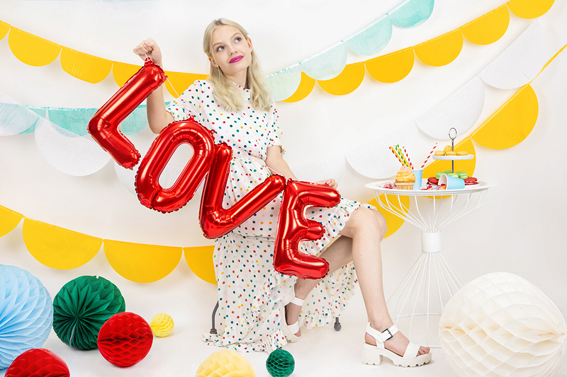 Mulher com vestido branco de bolinhas segurando balões vermelhos com a palavra LOVE numa decoração colorida