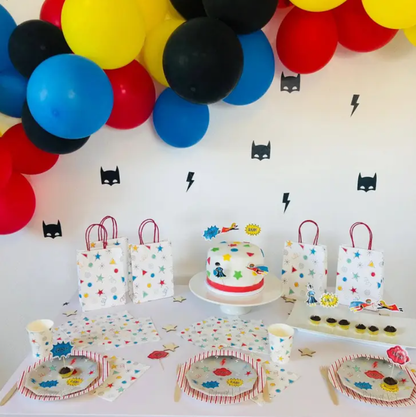 Mesa de festa infantil com bolo e decoração colorida de estrelas, balões coloridos e adesivos de morcego na parede