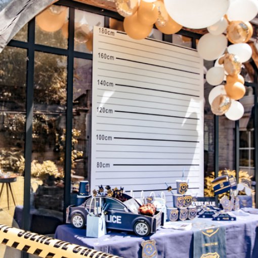 Decoração temática policial com viatura de brinquedo, balões brancos e dourados e painel de altura em centímetros.