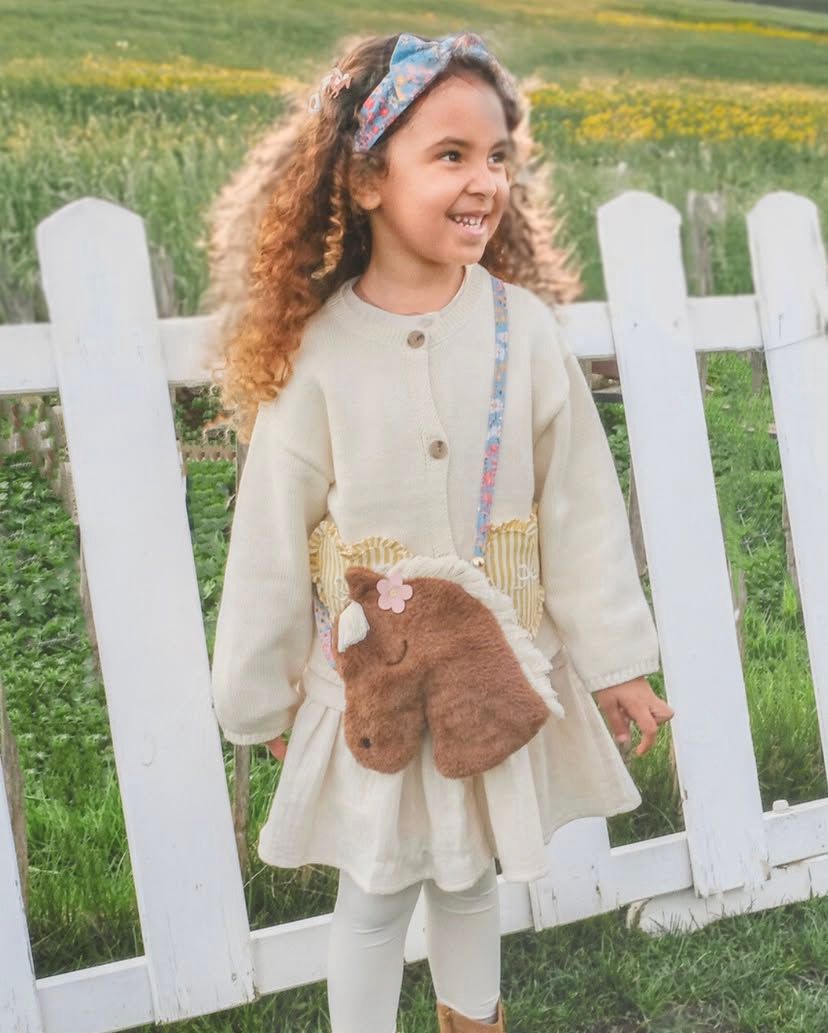 Menina com casaco bege e mala em forma de cavalo peludo castanho, com bandana azul, ao ar livre junto a cerca branca.