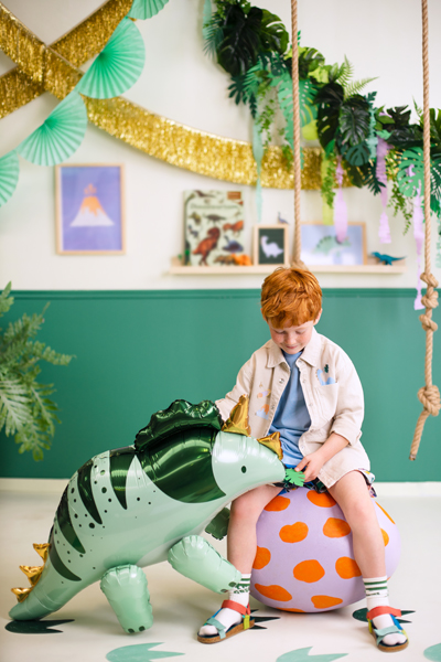 Menino sentado numa bola rosa com manchas laranja, balão verde com pattern de dinossauro, fundo decorado com folhas, faixas douradas e verdes e quadros