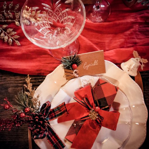 Mesa posta com pratos brancos, guardanapo vermelho com laço xadrez, cartão com o nome Cyrielle e taça de cristal em fundo vermelho