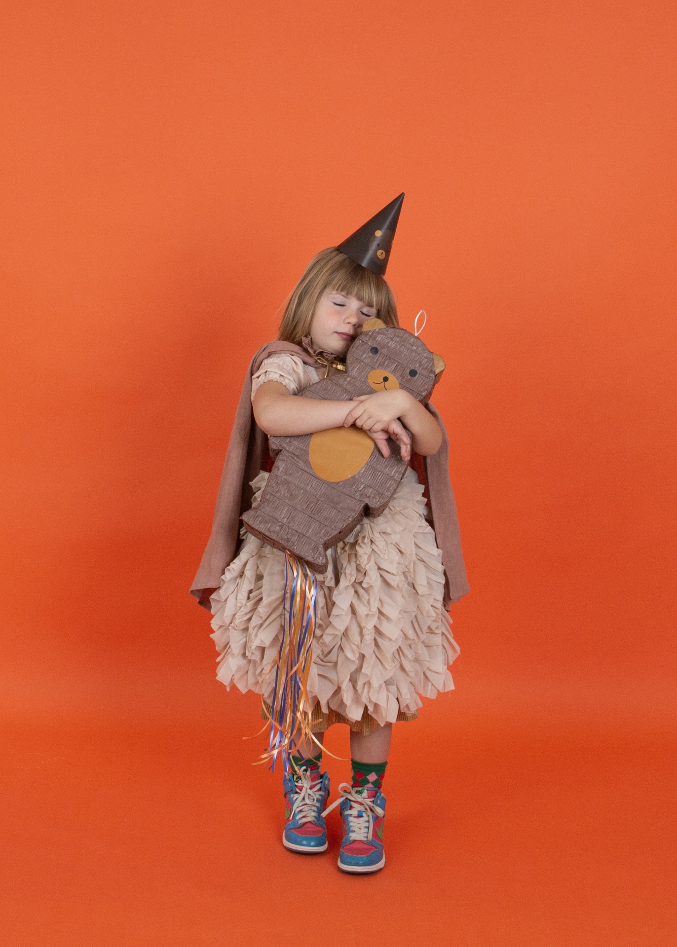 Menina com vestido volumoso bege e capa castanha segura peluche castanho em fundo laranja