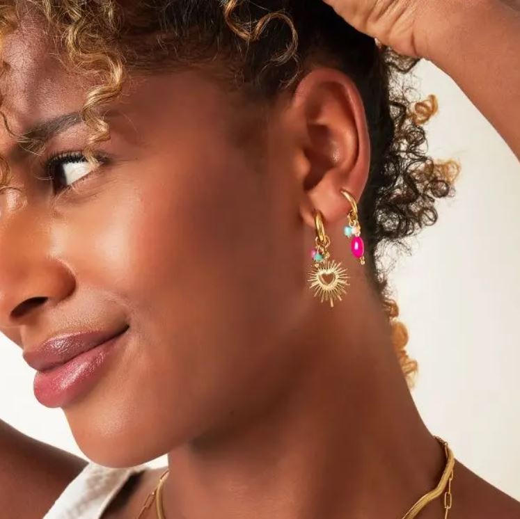 Mulher com brincos dourados, um em forma de sol e outro colorido, fundo branco