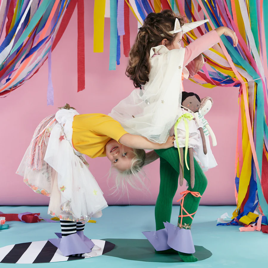 Crianças com fatos de unicórnio coloridos e fitas decorativas num ambiente lúdico.