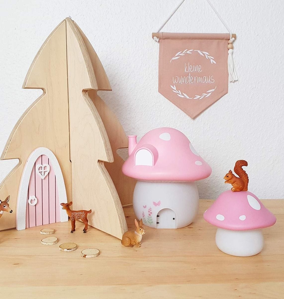 Decoração infantil com casa de madeira em forma de árvore, casas cogumelo cor de rosa, animais miniatura e tecido decorativo com texto em alemão.
