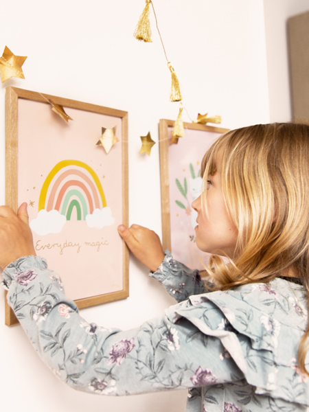 Menina loira a pendurar moldura com arco-íris e texto 'Everyday magic' num ambiente decorado com estrelas douradas