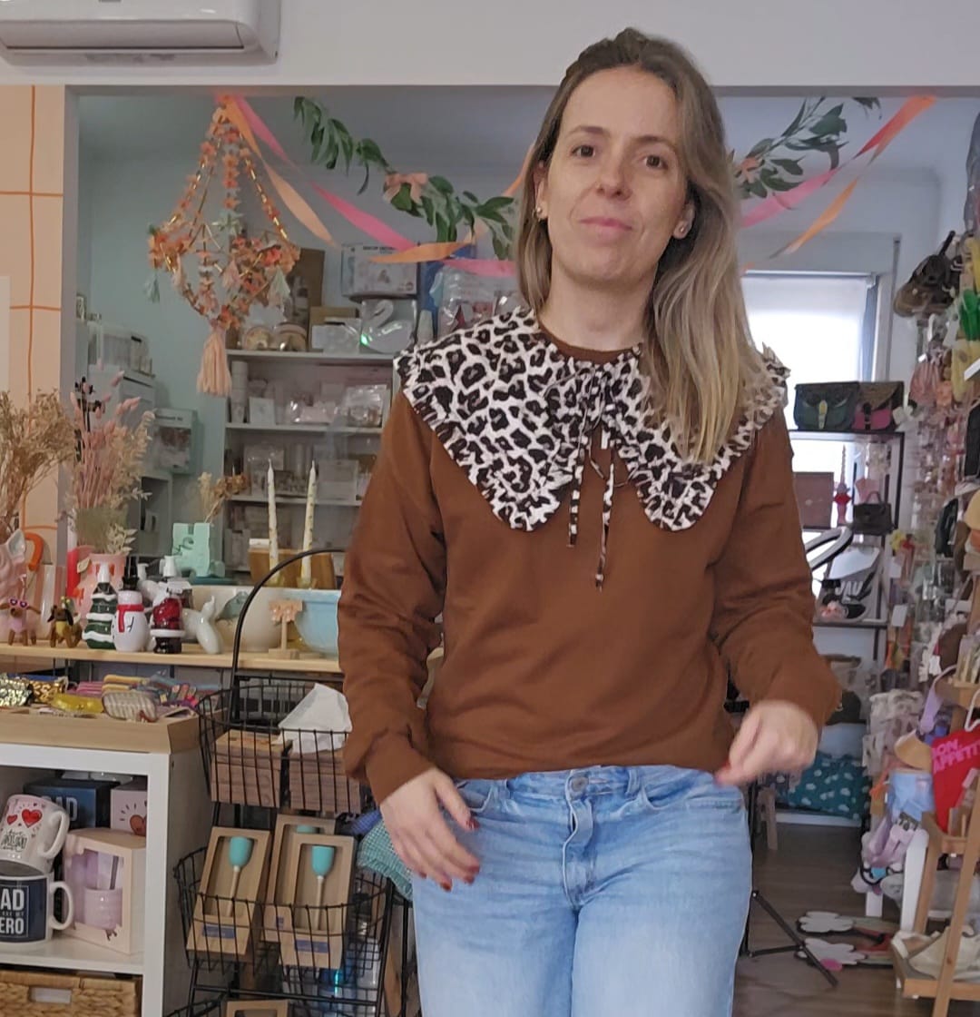 Mulher com camisola castanha e gola animal print numa loja com decoração e objetos diversos.