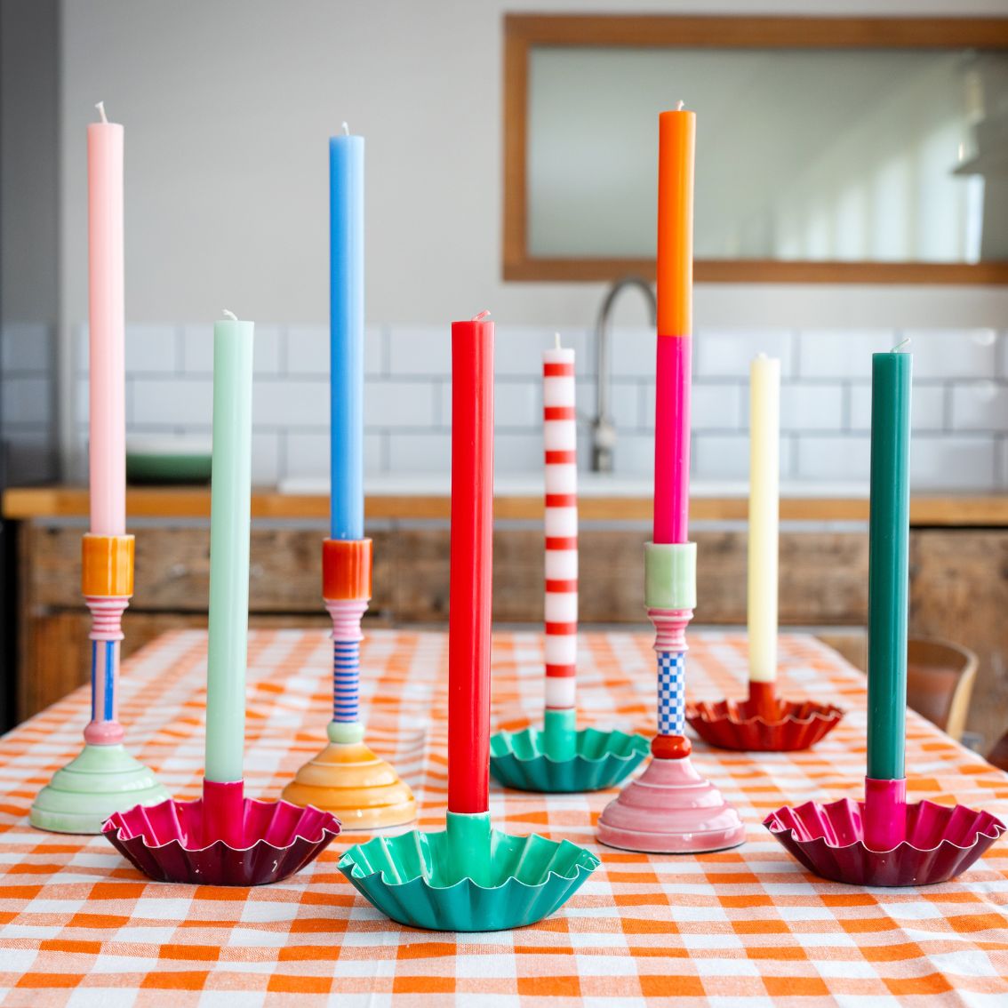 Sete castiçais coloridos com velas em cima de mesa quadriculada numa cozinha