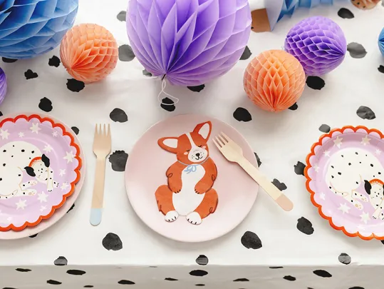 Pratos e talheres infantis com desenhos de animais e esferas de papel coloridas sobre toalha branca com manchas pretas