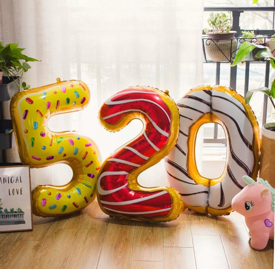 Balões dourados em forma dos números 5, 2 e 0 com padrões coloridos, numa sala com chão em madeira e luz natural.