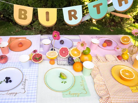 Mesa decorada para festa de aniversário com pratos coloridos, frutas e decoração, com faixa BIRTH ao fundo