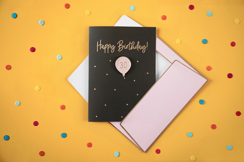 Cartão de aniversário preto com texto dourado e balão rosa ao centro, envelope branco e rosa, fundo amarelo com confetes coloridos.