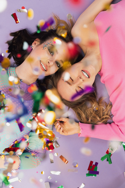Duas jovens deitadas em fundo lilás com confetes coloridos a cair