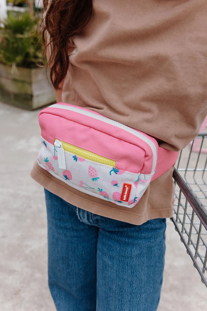 Bolsa de cintura rosa e branca com padrão de frutas, pessoa com t-shirt castanha e calças de ganga azuis junto a carrinho de compras