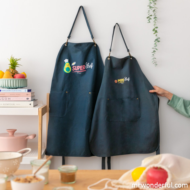 Dois aventais de tecido azul escuro com inscrições coloridas pendurados numa parede branca.