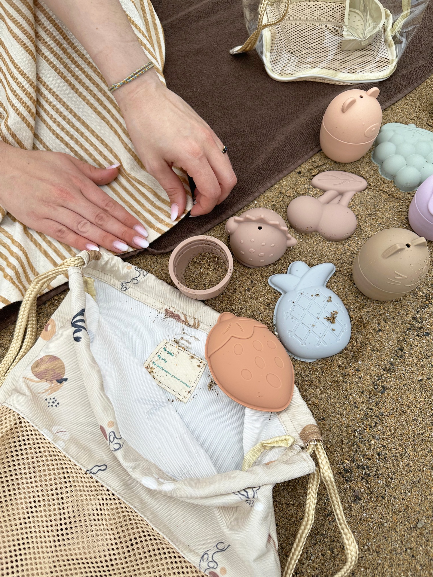 Brinquedos de silicone coloridos e sacos sobre areia com mãos femininas numa toalha listrada