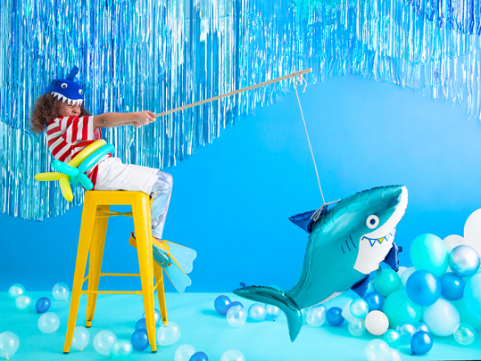 Criança com chapéu e boia em forma de tubarão, sentada num banco amarelo, segurando um balão de tubarão com vara de pesca num cenário azul decorado com balões.