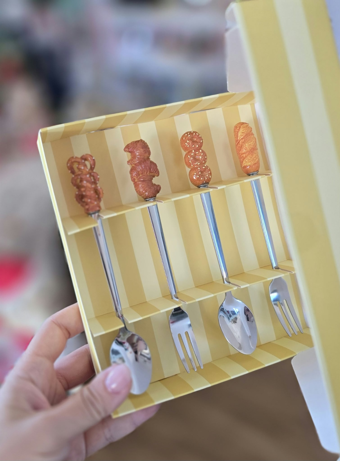 Talheres com cabos decorados com miniaturas de pão e pretzel numa caixa amarela às riscas.