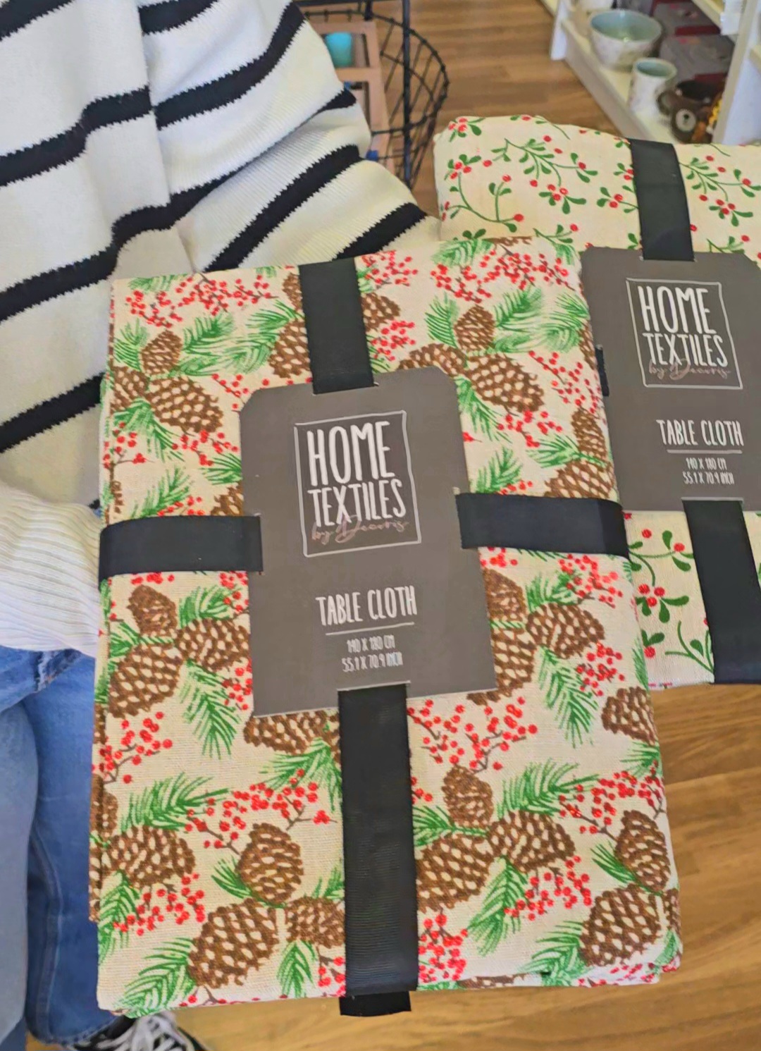 Toalha de mesa com padrão de pinhas, folhas e bagas embaladas com etiqueta preta.