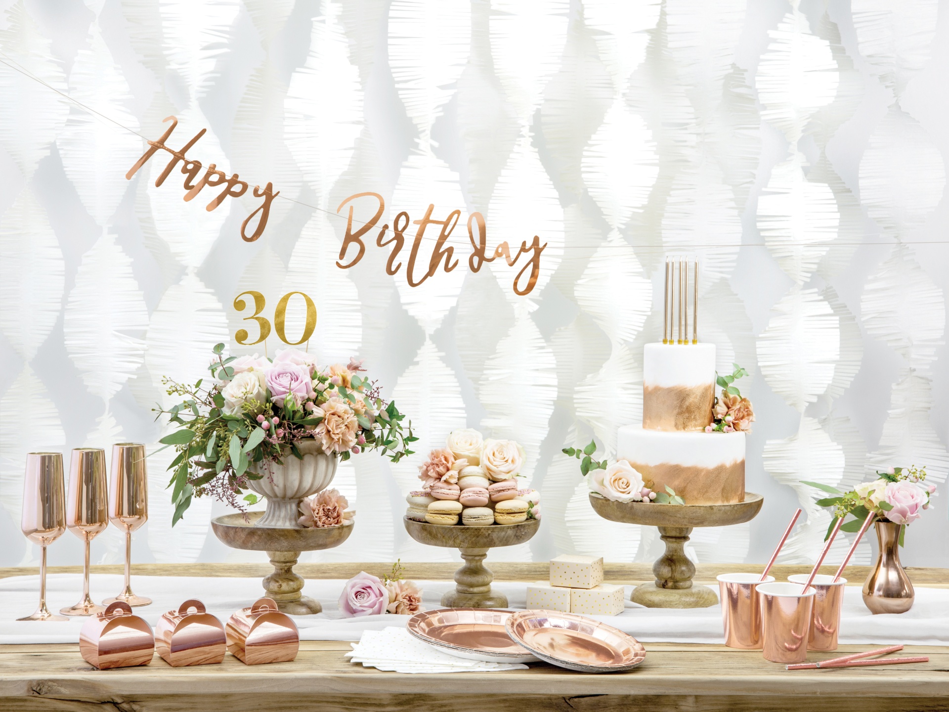 Mesa de aniversário decorada com bolo, cupcakes, arranjos florais, copos dourados e balões com texto 