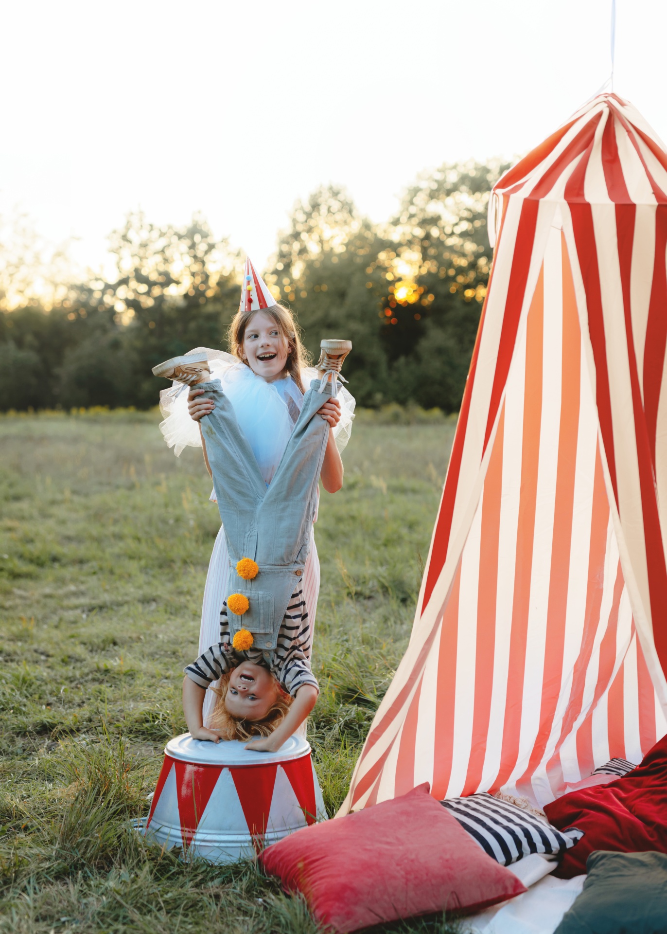 Criança a fazer parada de mãos numa lata junto de outra criança com chapéu de festa perto de tenda listrada
