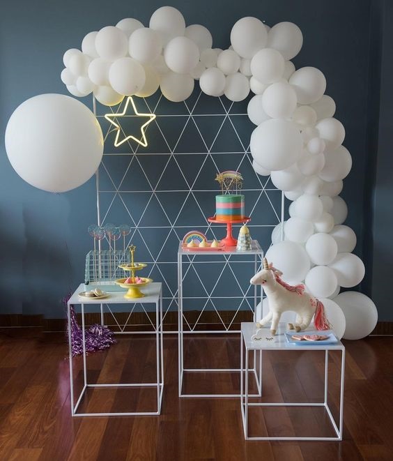 Decoração de festa com balões brancos em arco e mesa com bolo e unicórnio
