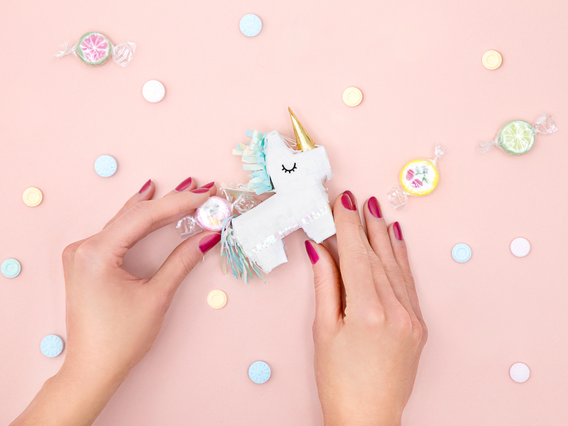 Mãos segurando piñata de unicórnio branco com fundo rosa e balas coloridas