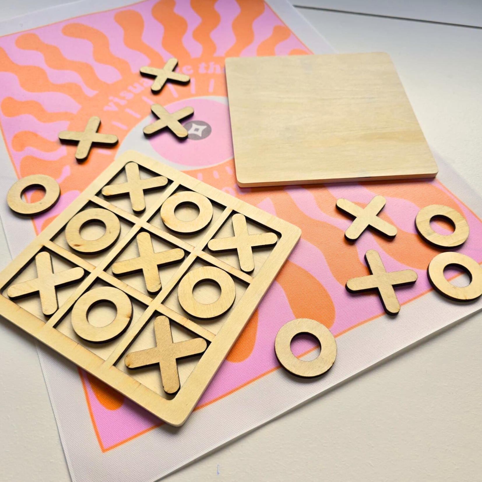 Jogo da velha em madeira clara com tabuleiro e peças X e O, sobre pano colorido