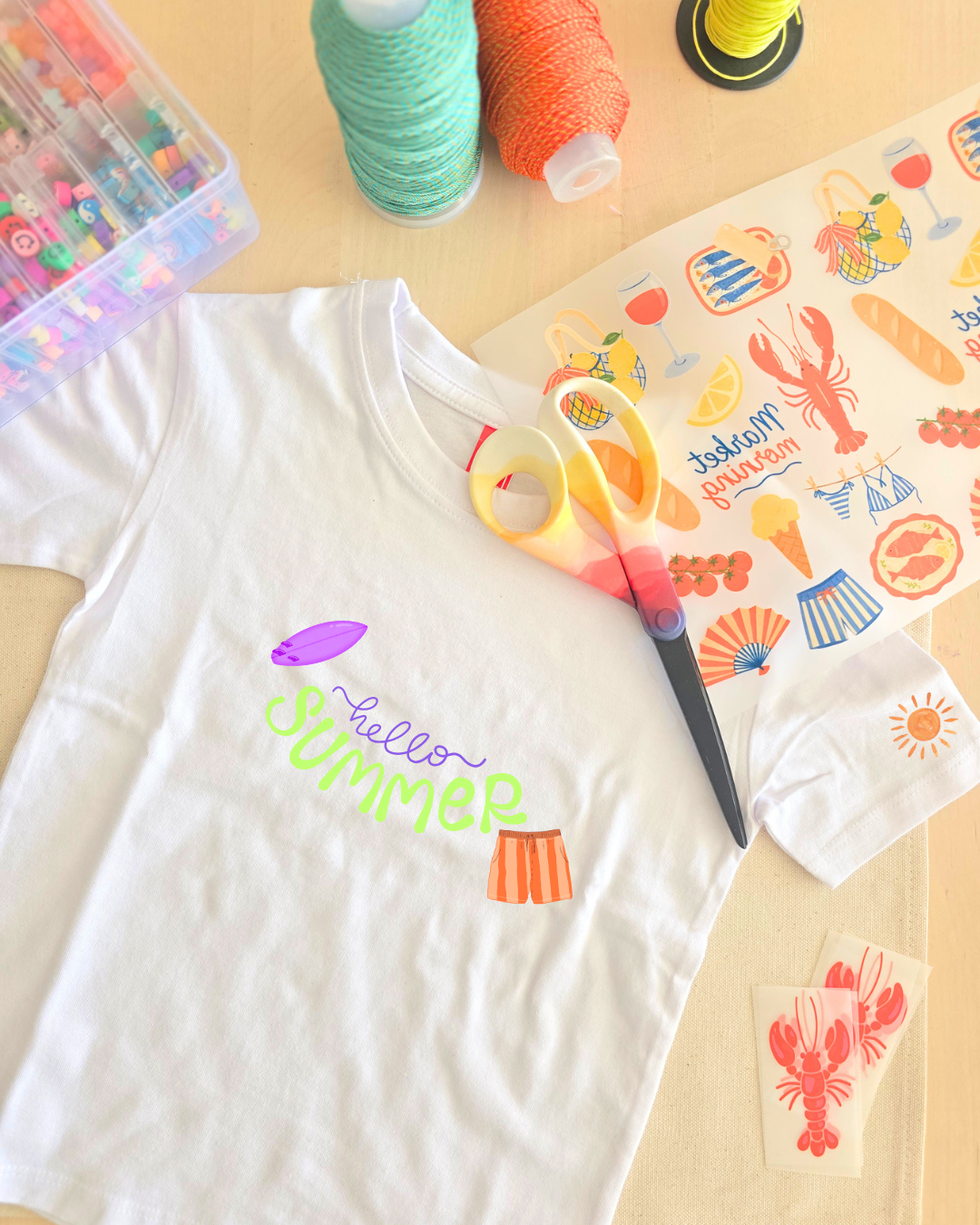 T-shirt branca com estampa sommer, tesoura colorida, rolos de linha e adesivos em mesa de madeira.