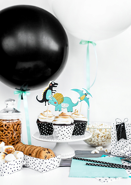 Mesa decorada com cupcakes com toppers de dinossauros, balões preto e branco, biscoitos e petiscos em caixas brancas com bolinhas pretas.