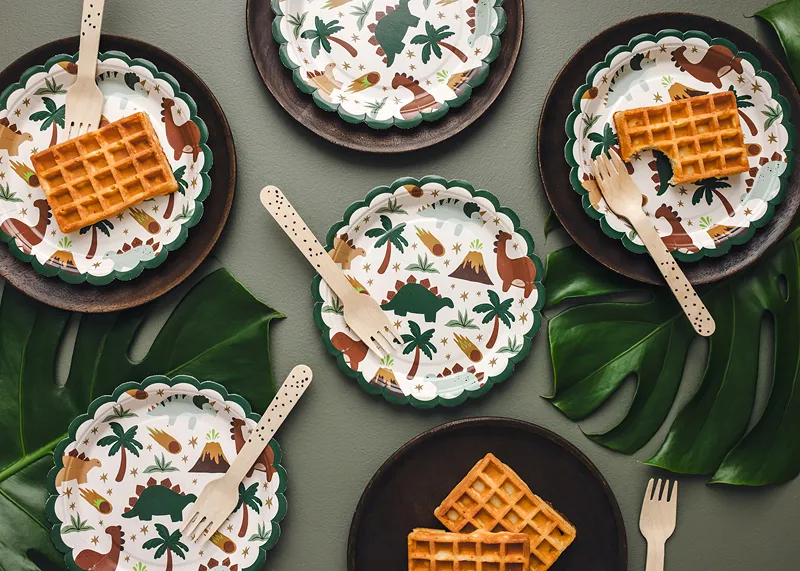 Pratos de papel com padrão de dinossauros a servir waffles com garfos de madeira sobre folhas verdes