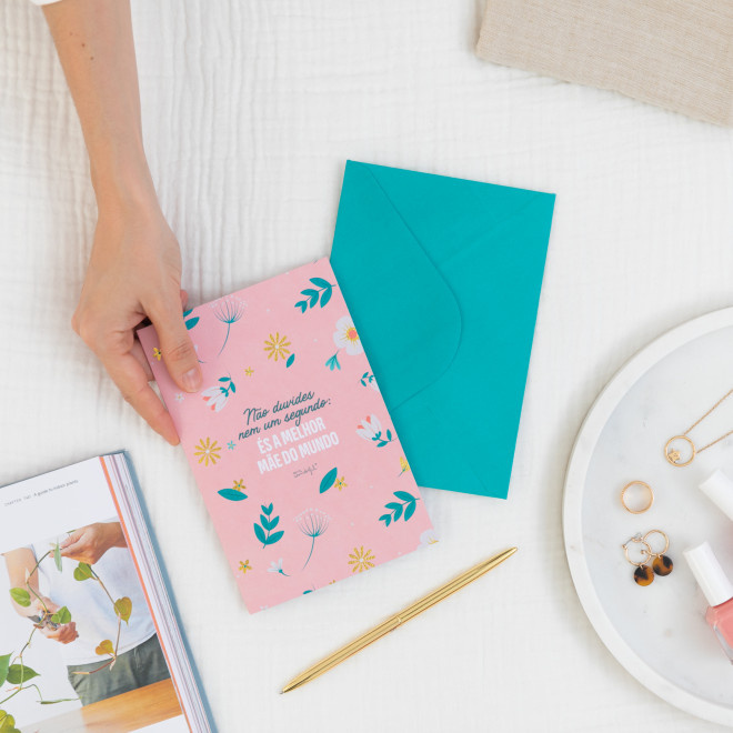 Cartão cor-de-rosa floral com mensagem, envelope azul, agulha dourada, revista, joias e vernizes sobre fundo branco.