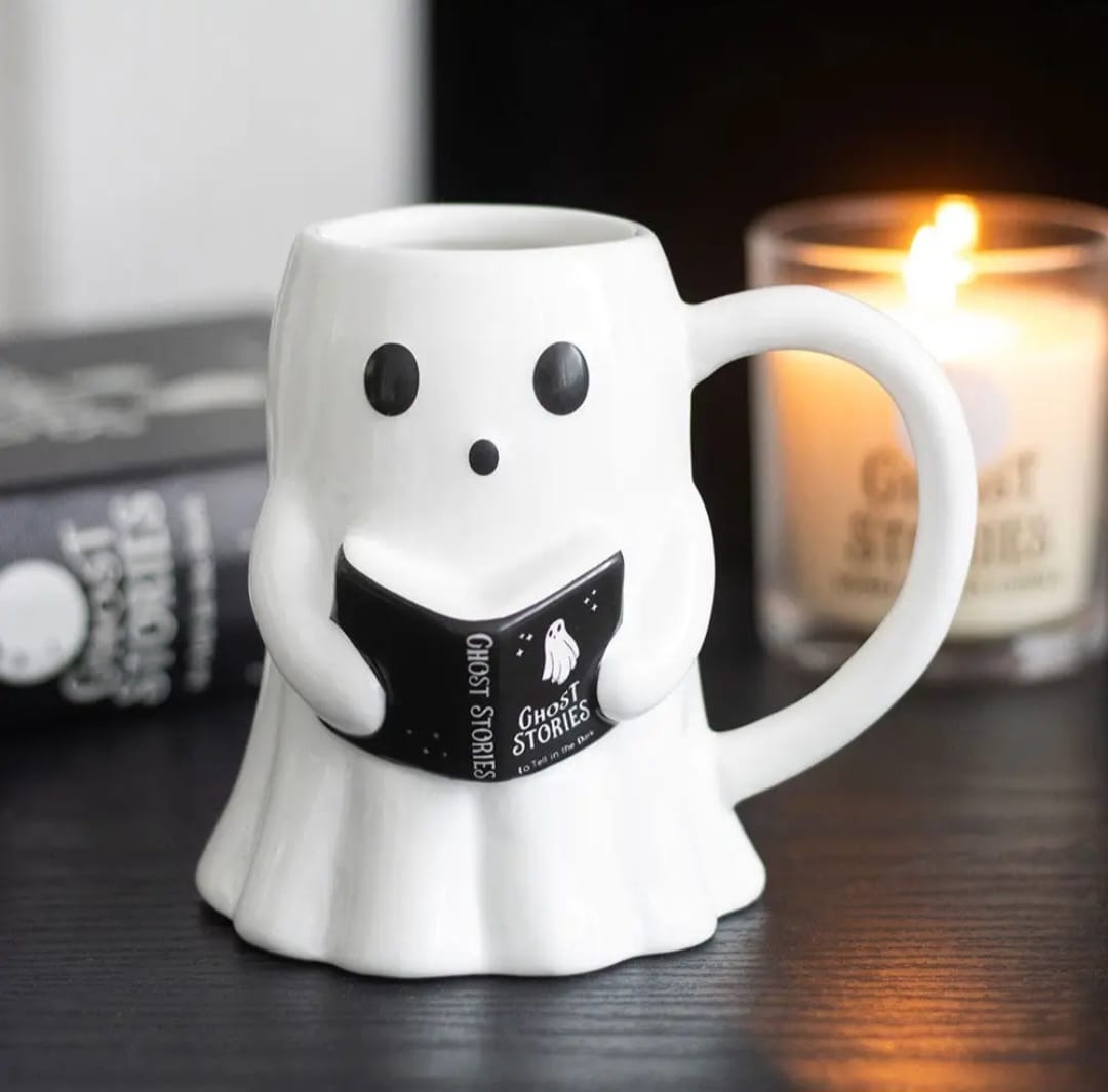 Caneca branca em forma de fantasma a segurar um livro preto com texto, numa mesa preta com vela acesa e livro ao fundo.