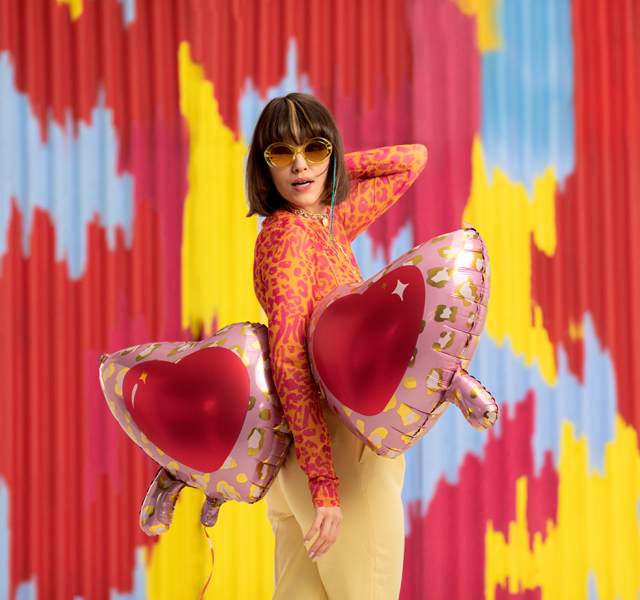 Mulher com blusa rosa de manchas vermelhas e calças amarelas, óculos escuros amarelos, segurando balões em forma de coração dourados e vermelhos, parede colorida ao fundo.