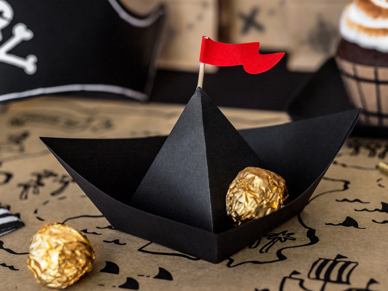 Barco de papel preto com bandeira vermelha e chocolates embrulhados em papel dourado sobre mesa com padrão de mapas piratas