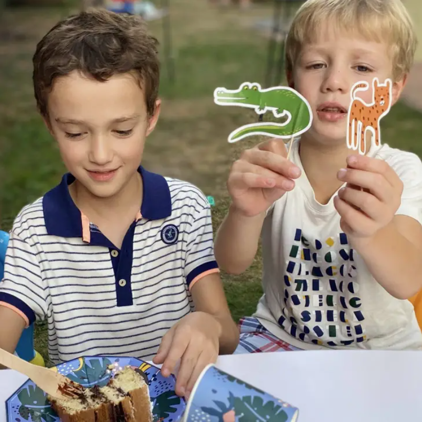 Duas crianças seguram recortes de papel de animais junto a um bolo e copo com padrões coloridos numa mesa ao ar livre