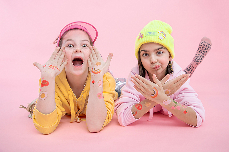 Duas raparigas com roupa casual colorida e desenhos coloridos nas mãos e braços deitadas em fundo rosa