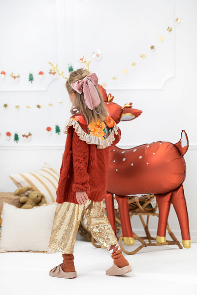 Menina com roupa de inverno colorida ao lado de balão inflável de veado metálico.