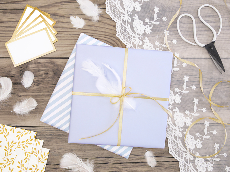 Presente embrulhado em papel branco com fita dourada e pena branca sobre madeira clara