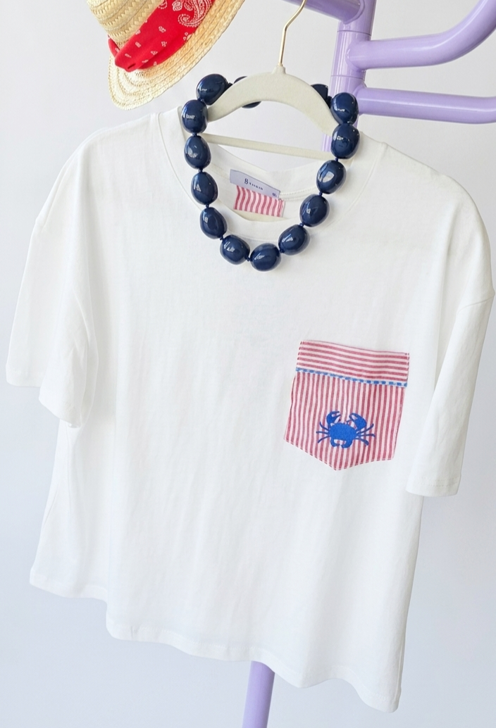T-shirt branca com bolso vermelho listrado e caranguejo azul pendurada com colar azul e chapéu de palha com lenço vermelho