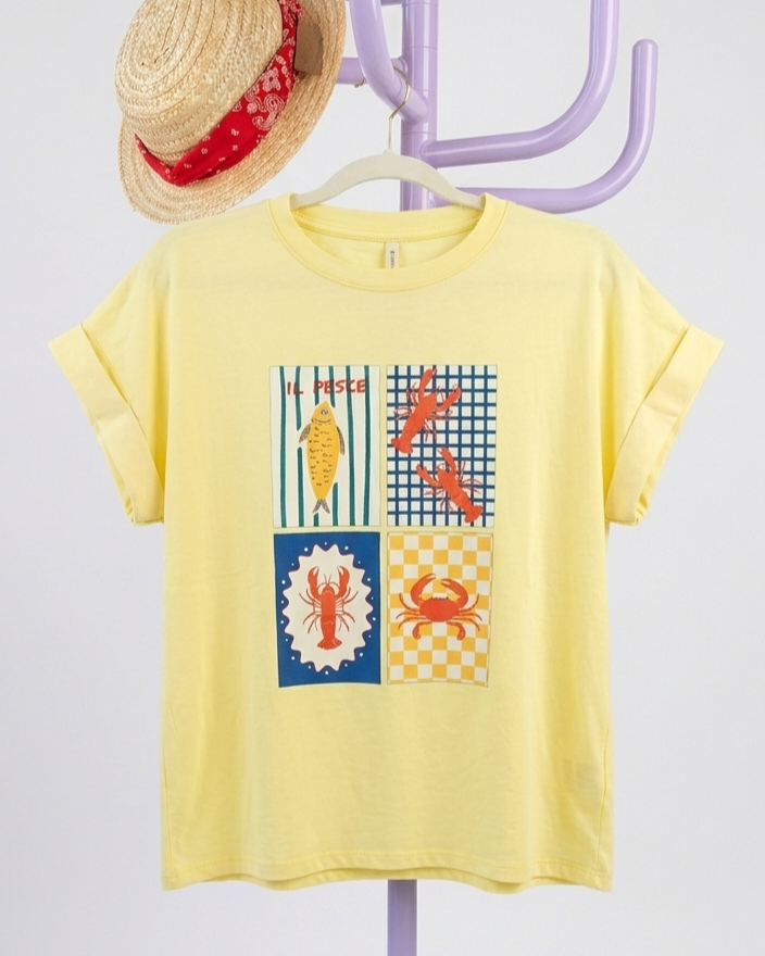 T-shirt amarela com estampas de peixe e crustáceos pendurada num cabideiro com chapéu de palha.