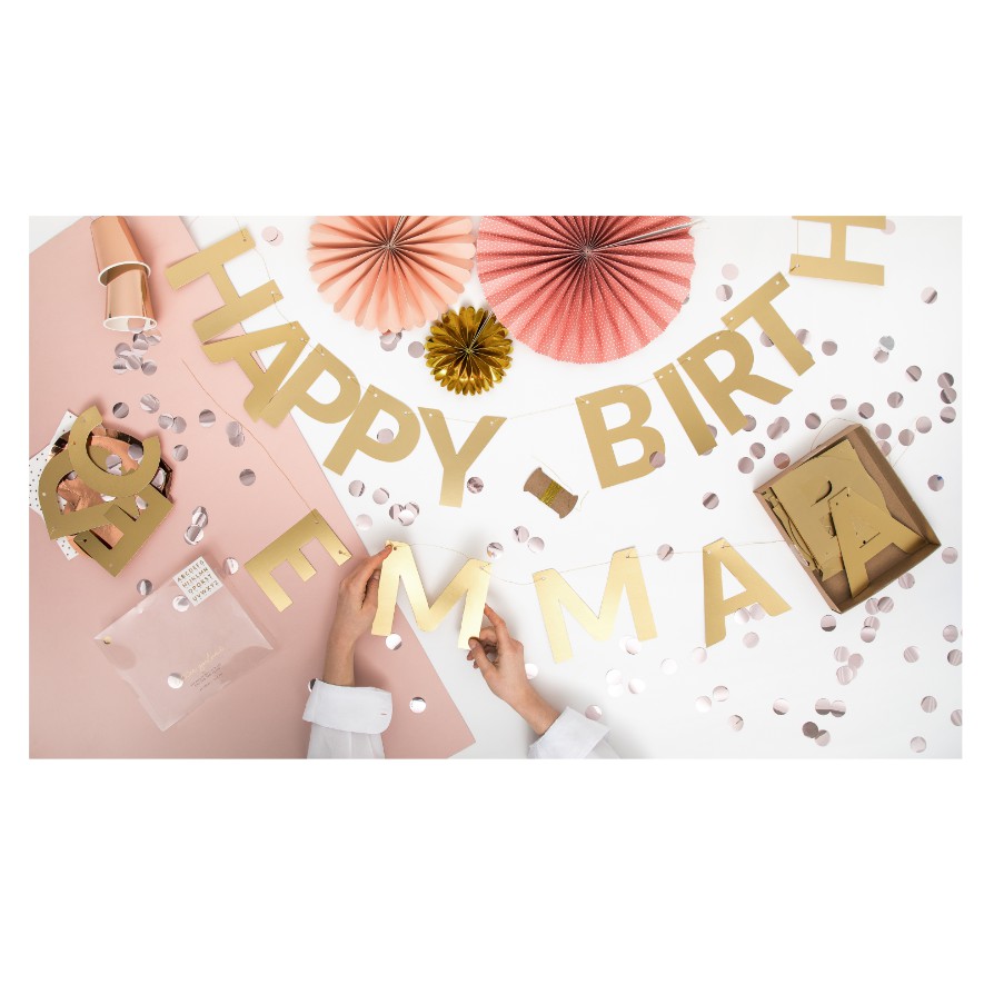 Decoração festiva com letras douradas, papel colorido rosa e dourado, confetes e duas mãos ajustando a decoração