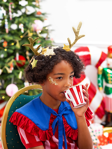 Criança com couraça dourada de rena a beber em caneca listrada, com decoração natalícia ao fundo