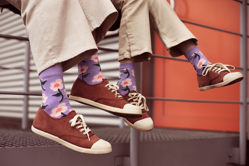 Ténis castanhos com meias roxas com flores, em ambiente industrial laranja.