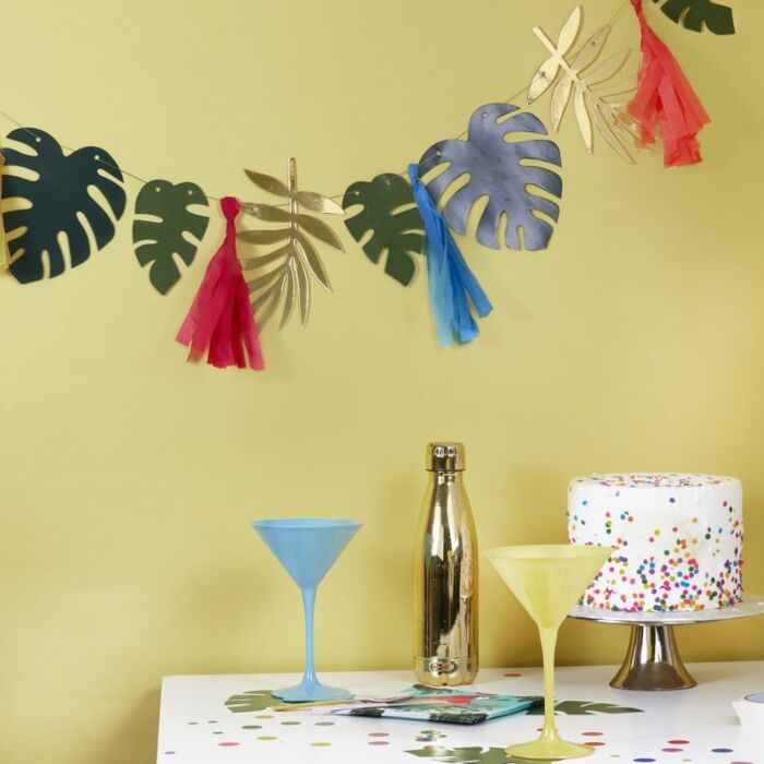 Decoração de festa com garland de folhas e tassels, garrafa metálica, copos de cocktail e bolo colorido